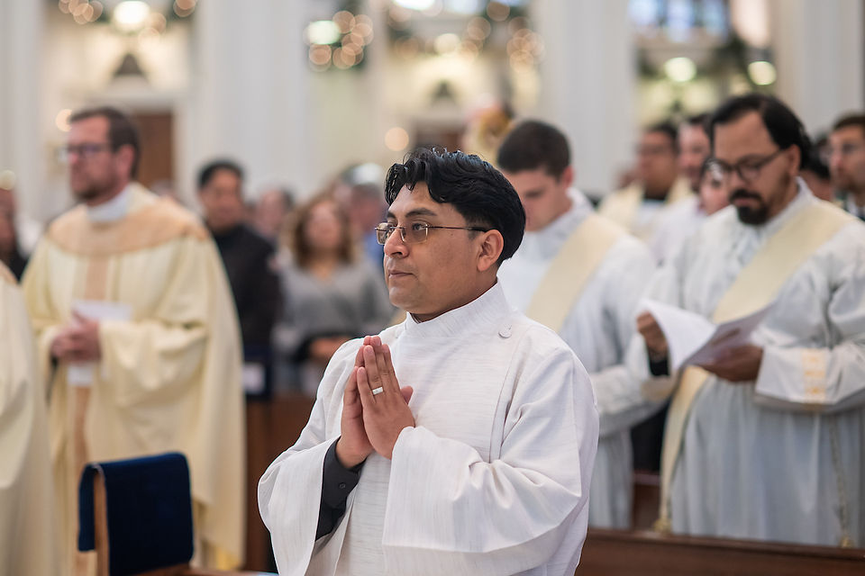A man in white robes prays in a church, standing with hands clasped. Others in similar attire hold papers. Soft lighting, focused ambiance.