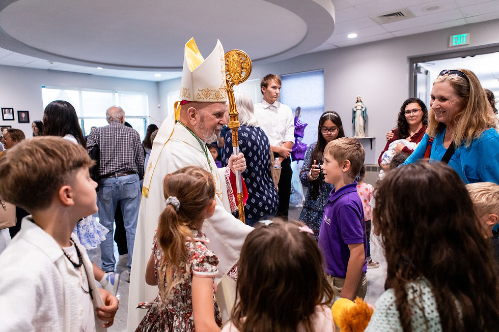 A bishop in ceremonial attire greets children in a church hall. Statues and people are visible in the background, creating a joyful atmosphere.