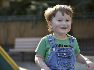 Smiling child in green shirt and denim overalls at playground, blurred bench and trees in background, sunlight creating a joyful mood.
