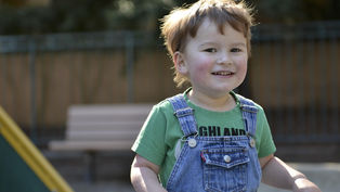 Smiling child in green shirt and denim overalls at playground, blurred bench and trees in background, sunlight creating a joyful mood.