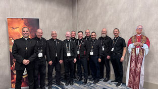 Ten men in black clerical attire smile for a group photo. A religious poster and cardboard figure are in the background. Indoor setting.