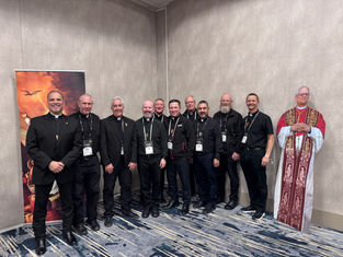 Ten men in black clerical attire smile for a group photo. A religious poster and cardboard figure are in the background. Indoor setting.