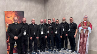 Ten men in black clerical attire smile for a group photo. A religious poster and cardboard figure are in the background. Indoor setting.