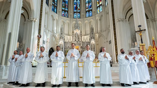 Twelve men in white robes with hands in prayer stand in a cathedral with stained glass windows and an ornate altar, creating a solemn mood.