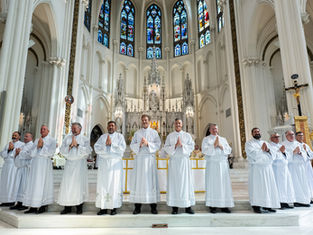 Twelve men in white robes with hands in prayer stand in a cathedral with stained glass windows and an ornate altar, creating a solemn mood.