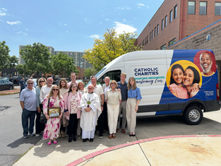 Group of people smiling in front of a van with "Catholic Charities" text. They're outdoors with a building and trees in the background.