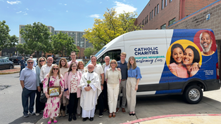 Group of people smiling in front of a van with "Catholic Charities" text. They're outdoors with a building and trees in the background.