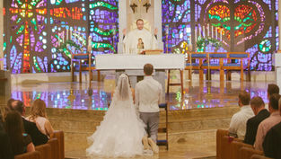 Bride and groom kneel at altar in a church with colorful stained glass backdrop. Priest stands with raised hands. Ceremony atmosphere.