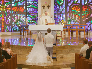 Bride and groom kneel at altar in a church with colorful stained glass backdrop. Priest stands with raised hands. Ceremony atmosphere.