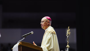 A bishop in ornate robes speaks at a podium with a microphone. He wears a pink zucchetto. Dark background with candle and staff visible.