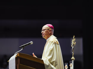 A bishop in ornate robes speaks at a podium with a microphone. He wears a pink zucchetto. Dark background with candle and staff visible.