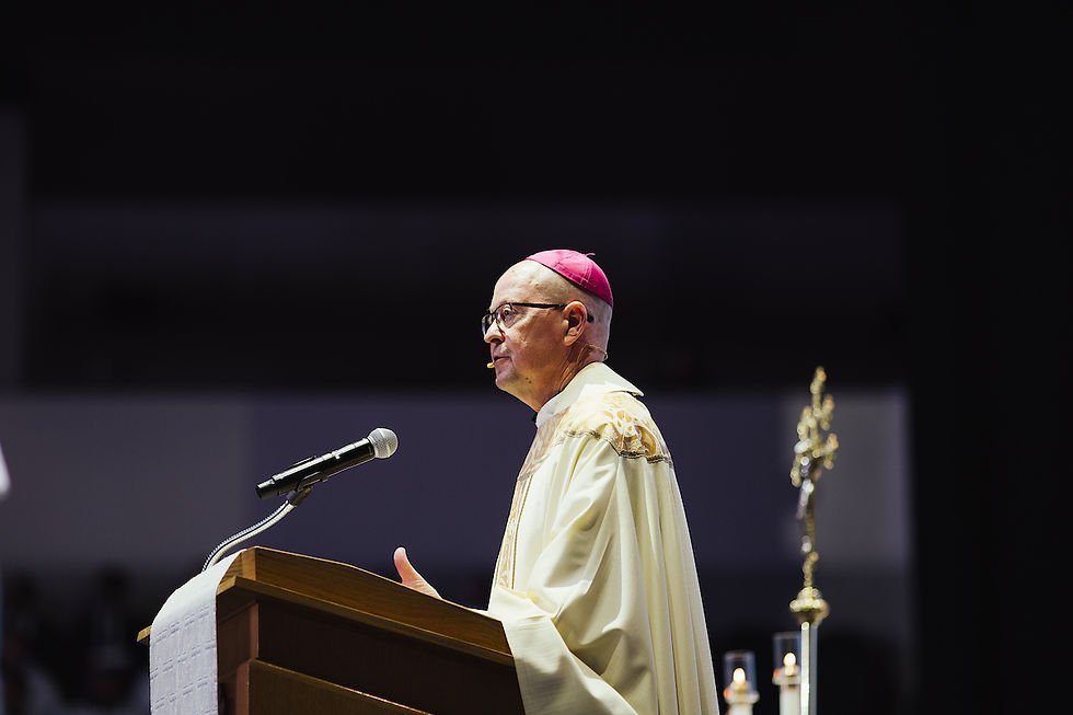 A bishop in ornate robes speaks at a podium with a microphone. He wears a pink zucchetto. Dark background with candle and staff visible.