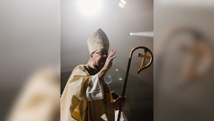 Man in ornate religious attire holds a staff with a cross, under bright light in a dim setting, gesturing with his hand.