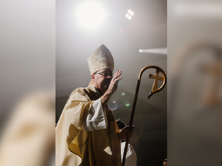 Man in ornate religious attire holds a staff with a cross, under bright light in a dim setting, gesturing with his hand.