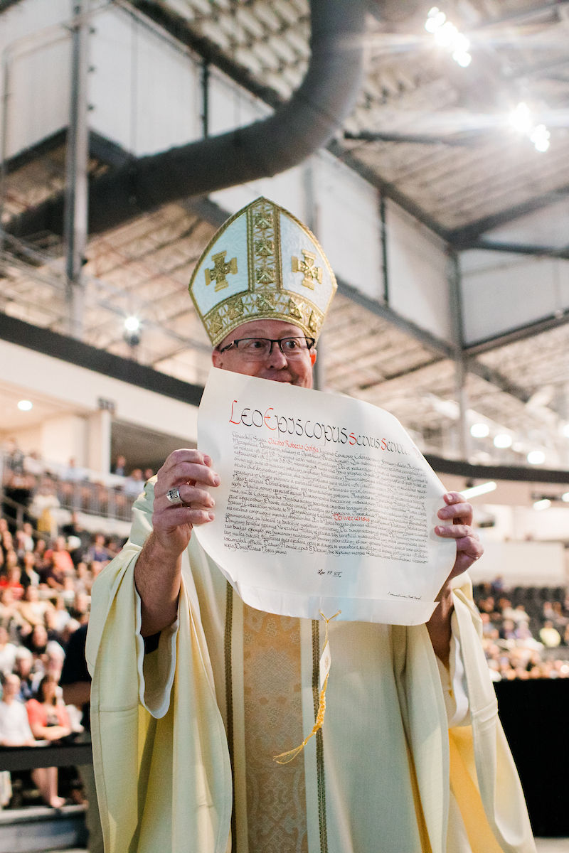 A newly installed bishop holds up an apostolic mandate as the congregation looks on.