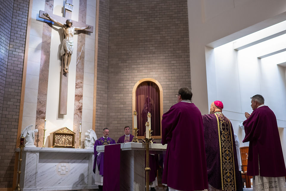 Clergy in purple vestments stand before an altar under a large crucifix in a church. The setting is solemn, with soft light filtering in.