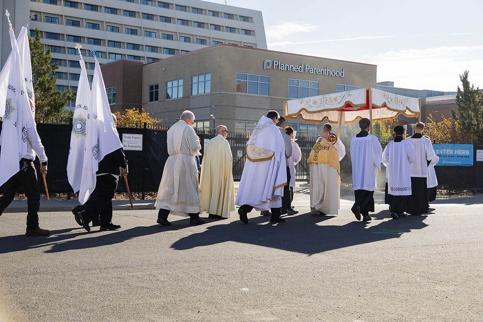 A religious procession in traditional robes passes a Planned Parenthood building. Participants carry banners and a canopy. Bright, sunny day.