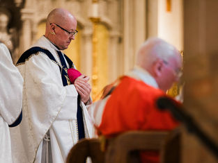 Clerics in ornate robes, one in red, another holding a pink hat, in a church setting with arched decor, conveying a solemn mood.