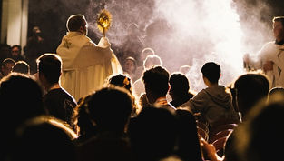 Priest holding a monstrance, surrounded by seated congregation. Smoke and dim lighting create a solemn, reverent atmosphere.