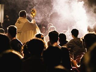 Priest holding a monstrance, surrounded by seated congregation. Smoke and dim lighting create a solemn, reverent atmosphere.