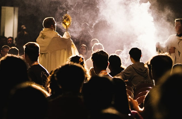 Priest holding a monstrance, surrounded by seated congregation. Smoke and dim lighting create a solemn, reverent atmosphere.