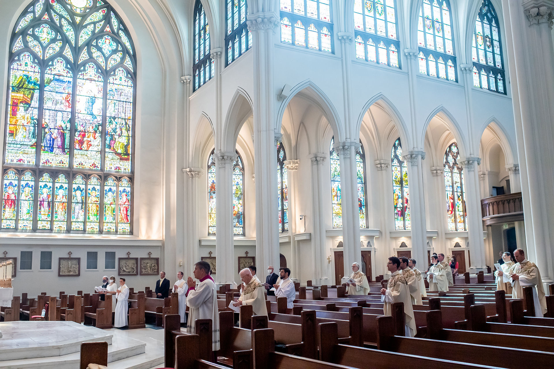 Priests and clergy stand in prayer inside a cathedral with grand stained glass windows and arched ceilings, evoking a solemn atmosphere.
