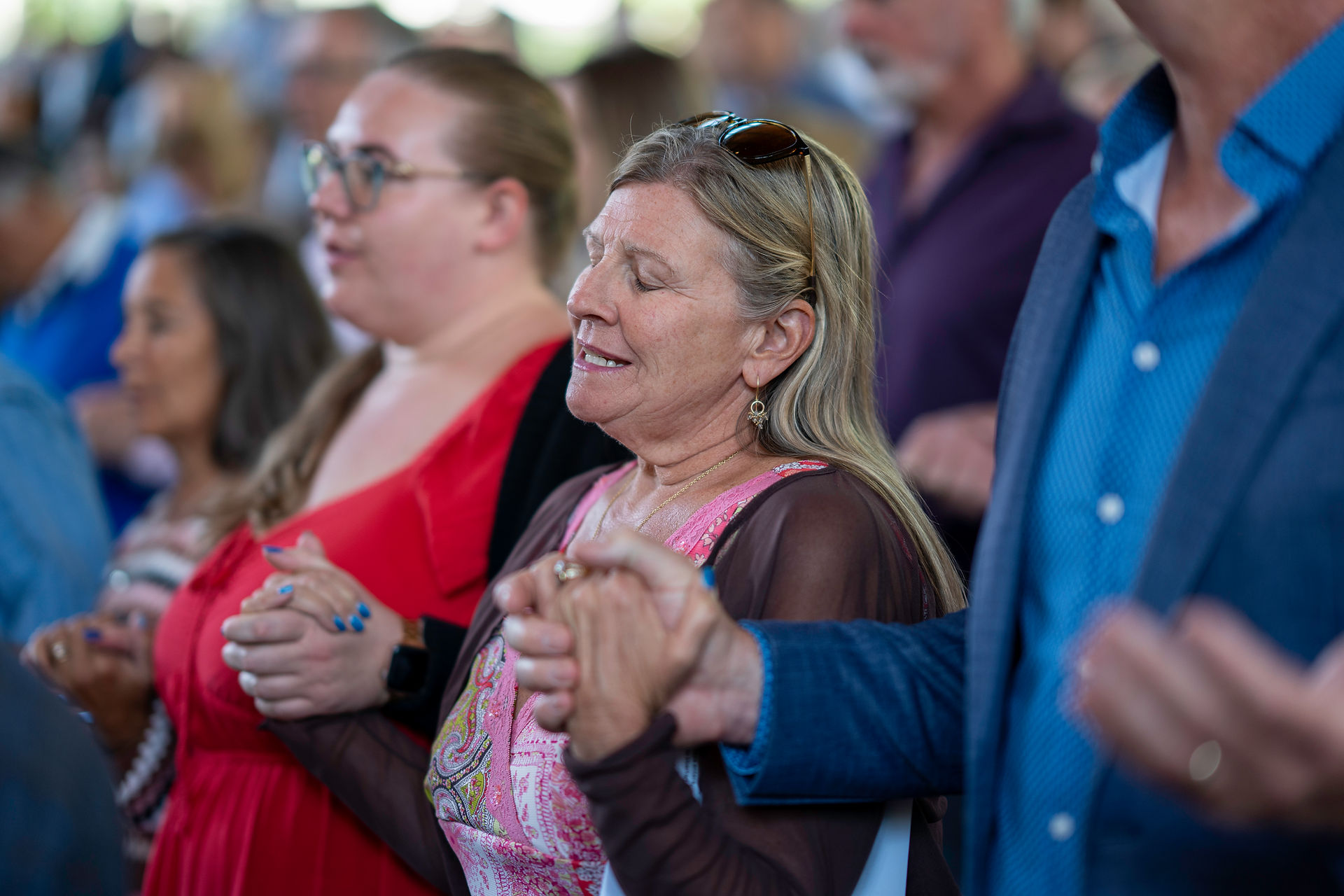 People holding hands in a group, eyes closed, in a serene setting. A woman in a pink dress stands out. The mood is peaceful.
