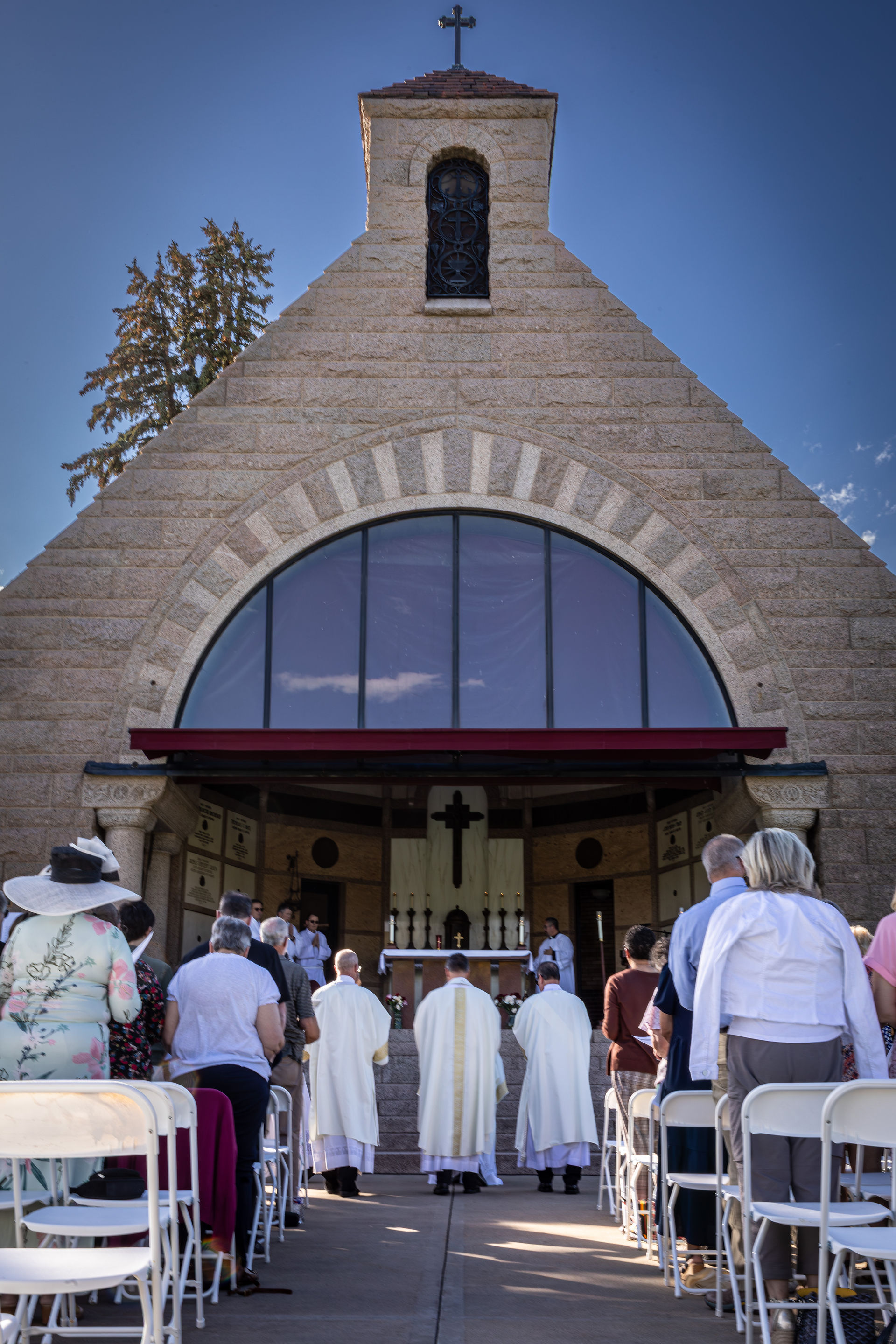People gather outside a stone church, with three clergy in white heading towards an altar, under a clear blue sky.