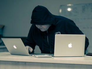 Hooded person working on two laptops at a desk, focused intently. Background shows a whiteboard. Notepad and pen on the desk.