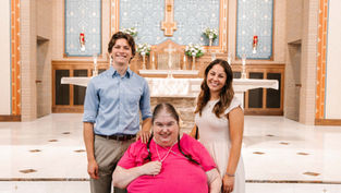 Three people smiling in a church setting. One person in a bright pink shirt seated; two others stand beside in casual attire. Bright, ornate backdrop.