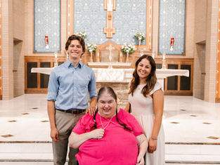Three people smiling in a church setting. One person in a bright pink shirt seated; two others stand beside in casual attire. Bright, ornate backdrop.
