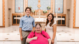 Three people smiling in a church setting. One person in a bright pink shirt seated; two others stand beside in casual attire. Bright, ornate backdrop.