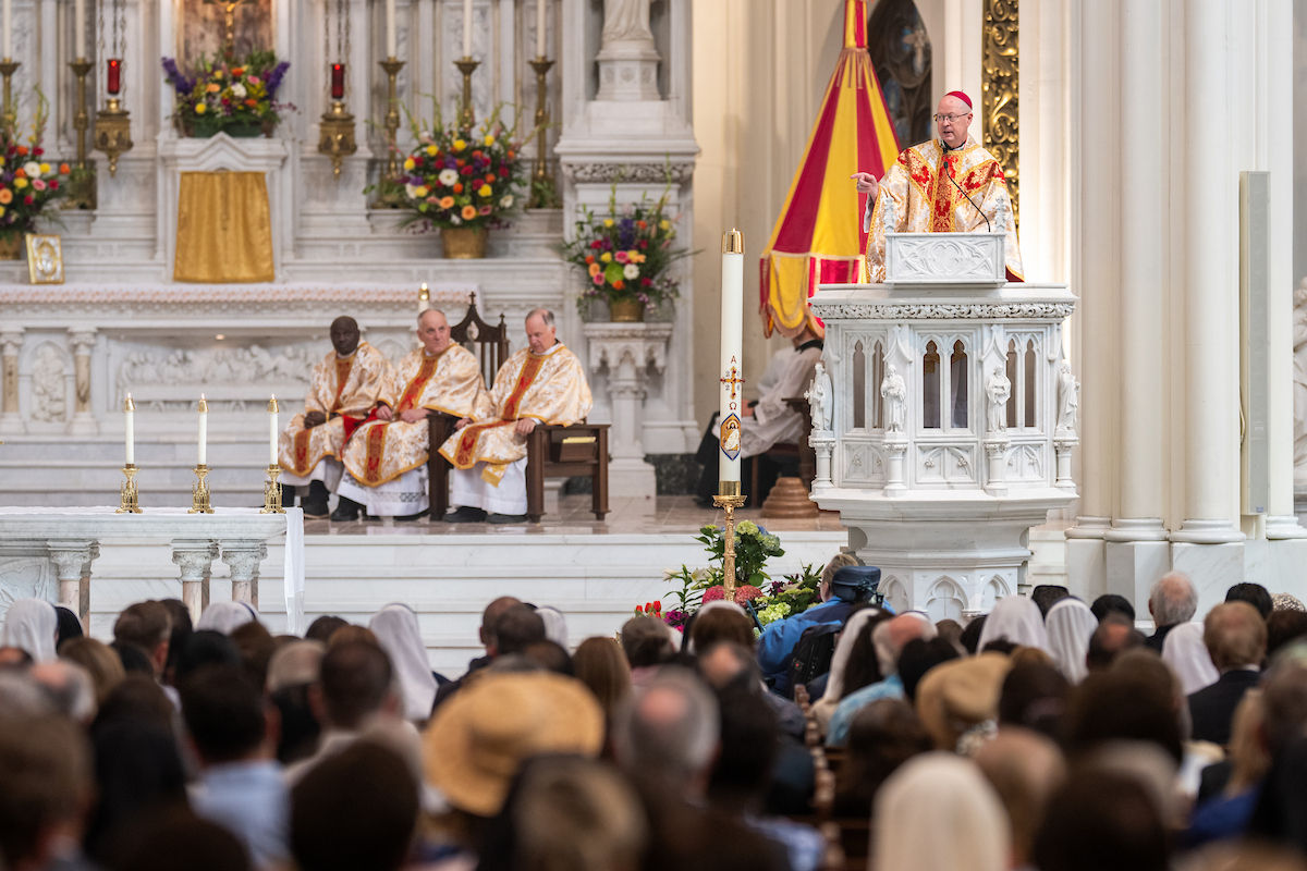 A bishop preaches from the pulpit during Mass as clergy sit behind him near the altar.