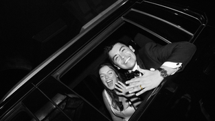 A smiling couple leans out of a car window at night, showing off rings. The man wears a tuxedo. Black and white image. Joyful mood.