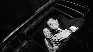 A smiling couple leans out of a car window at night, showing off rings. The man wears a tuxedo. Black and white image. Joyful mood.