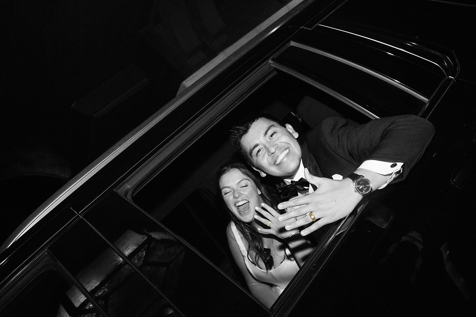 A smiling couple leans out of a car window at night, showing off rings. The man wears a tuxedo. Black and white image. Joyful mood.