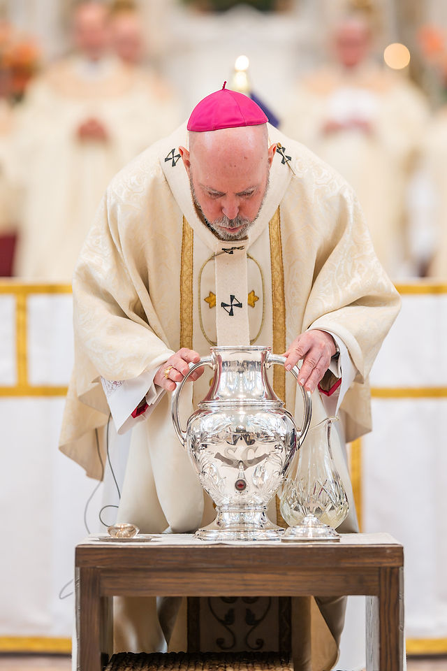 A bishop in ornate robes and a red cap bows over a large silver jug on a wooden table in a church setting, performing a ceremonial act.