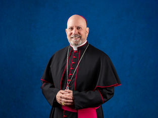 Cleric in black and red robes smiles, standing against a blue background. He wears a gold ring and chain, conveying a serene and formal mood.