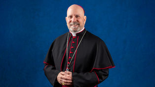 A smiling religious figure in black and red attire stands against a blue background, hands folded. Silver chain and ring visible.