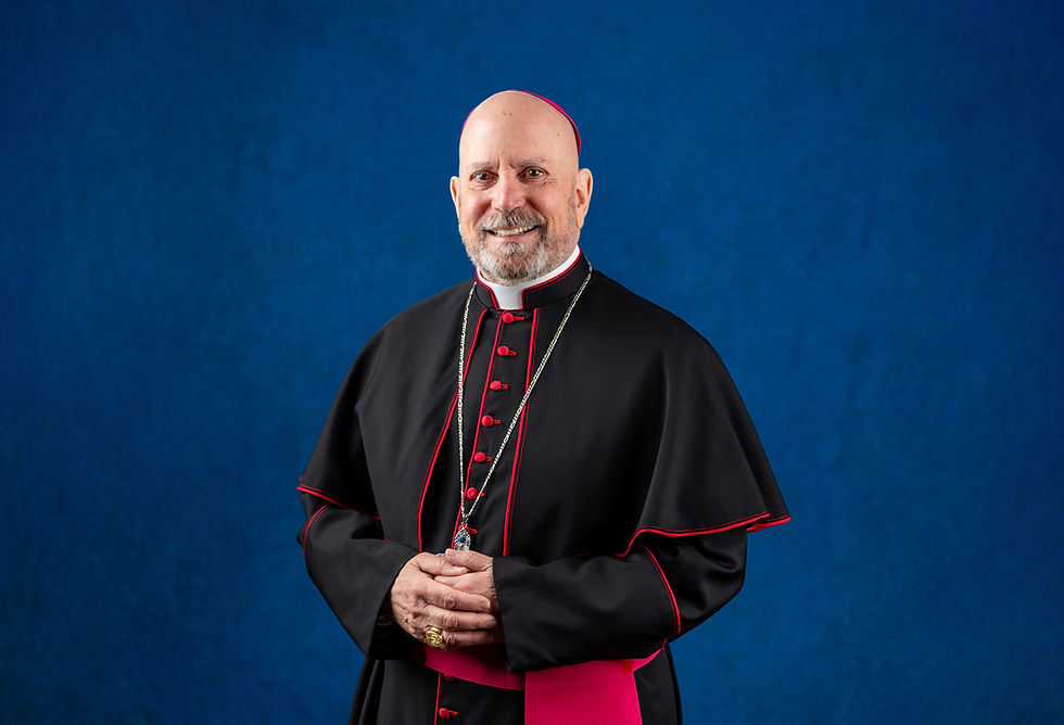 A smiling religious figure in black and red attire stands against a blue background, hands folded. Silver chain and ring visible.