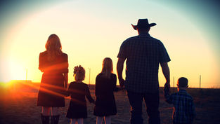 A family of five, holding hands, stands in silhouette against a vibrant sunset. They are in open country, with a peaceful, warm ambiance.