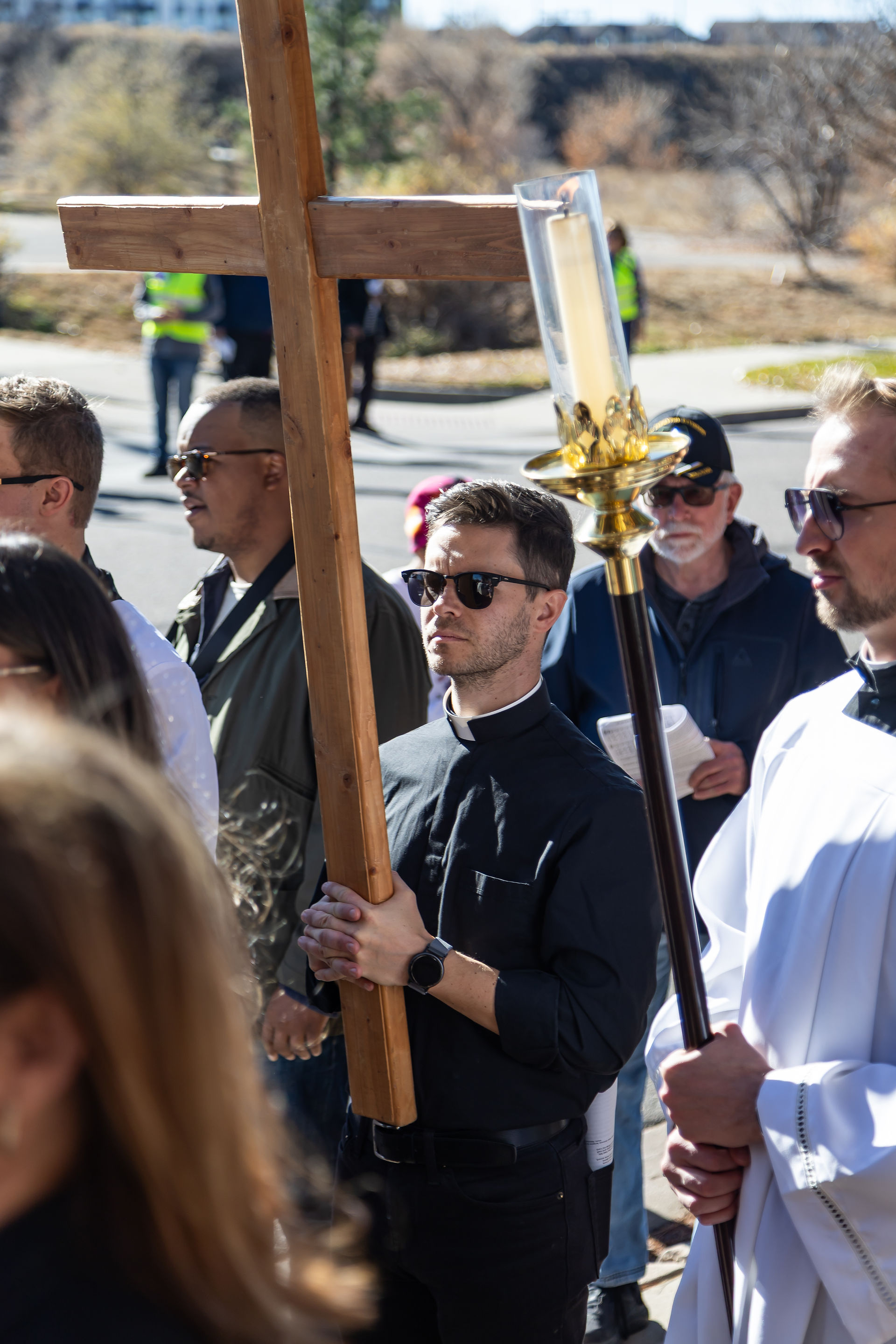 A procession with a man in black holding a wooden cross. Another holds a candle. Sunny day with people and trees in the background.
