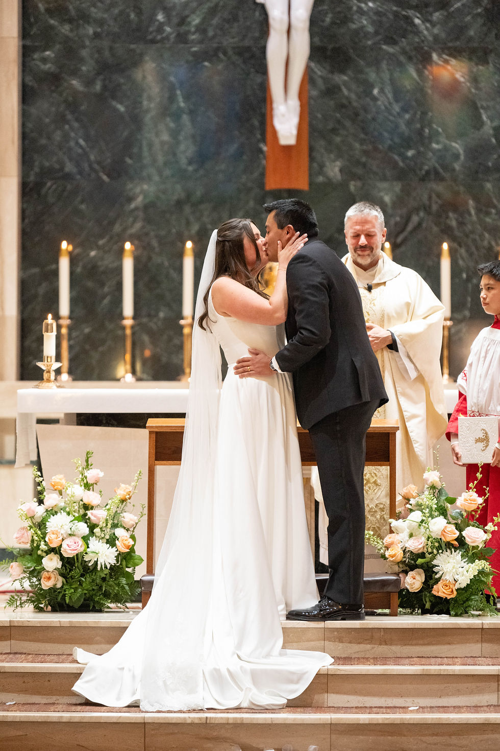 Bride and groom kiss at altar; priest blesses them. Candles lit, flowers around, serene mood in ornate church setting.