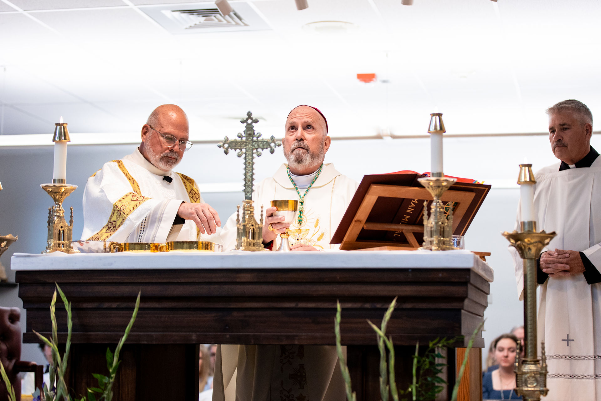 Clergy in white liturgical vestments stand at an altar holding a chalice. Candles, a cross, and a book are on the altar.