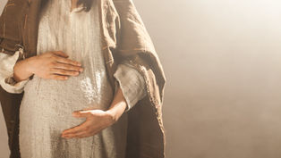 A person in a brown shawl and textured white dress cradles their pregnant belly. Soft warm lighting creates a peaceful mood.