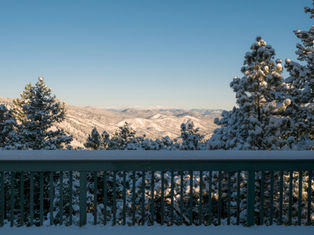 Snow-covered trees and hills viewed from a wooden deck railing under a clear blue sky. The scene is peaceful and serene.