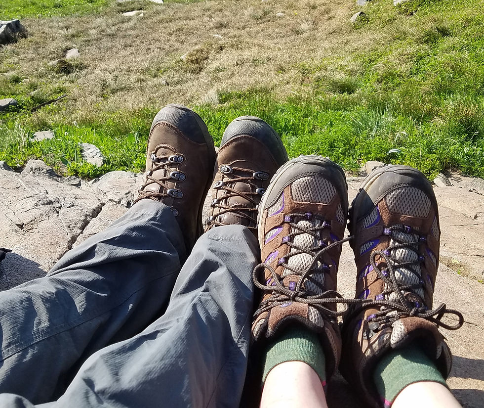 Two pairs of hiking boots resting on rocky ground, with greenery and dry grass in the background, suggesting a peaceful outdoor setting.