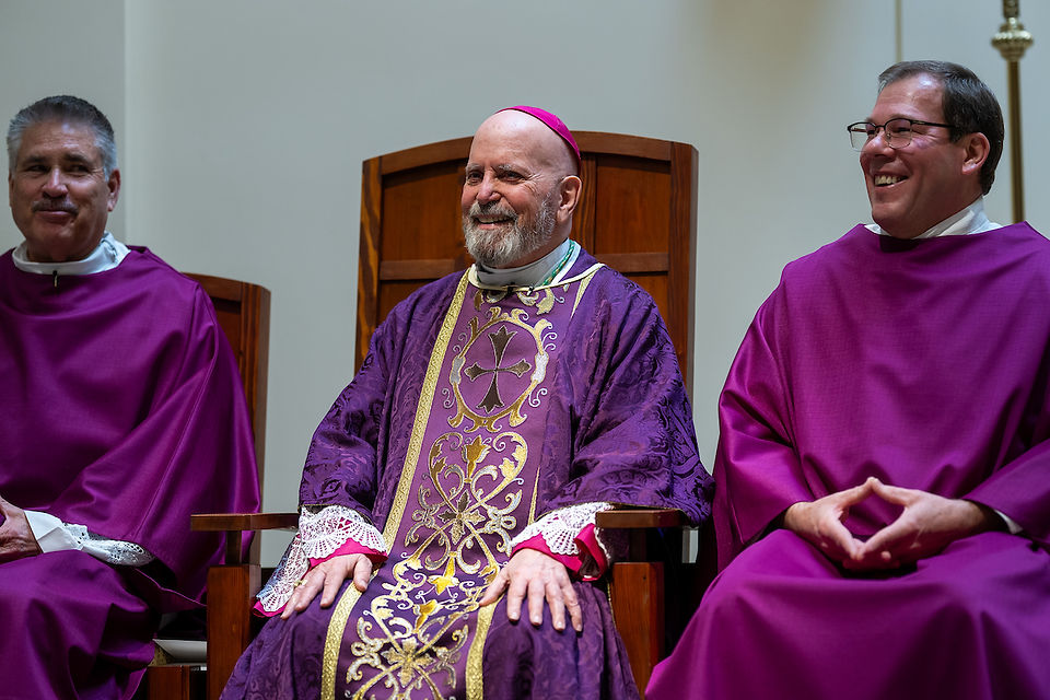 Three men in ornate purple robes sit on wooden chairs, smiling in a calm, bright setting. The central figure wears a decorated chasuble.