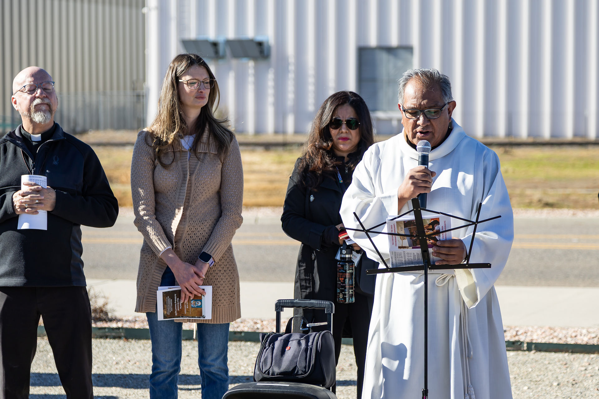 A group of people stands outside a building. A man in white robes speaks into a microphone, holding papers. Others listen, holding pamphlets.