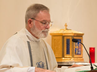 Priest in white robe reading at a lectern, with a microphone. Gold tabernacle and red candle in the background. Calm and focused mood.